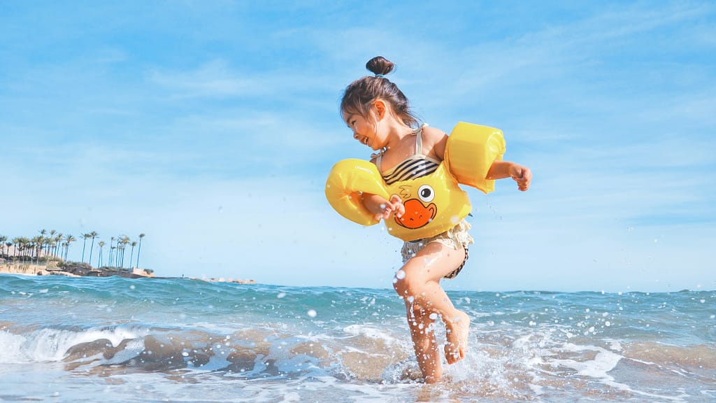 Image: A child wearing arm floaties and splashing in the water on the beach, under the beating sun in our changing climate.