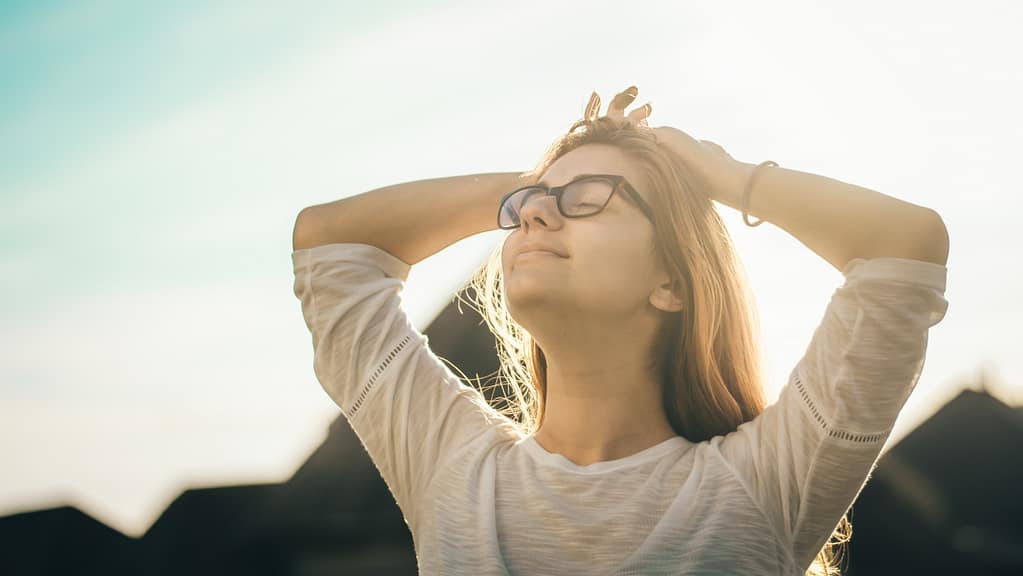 Image: A blonde woman wearing glasses, standing outside with her hands behind her head, with a look of ease and joy on her face
