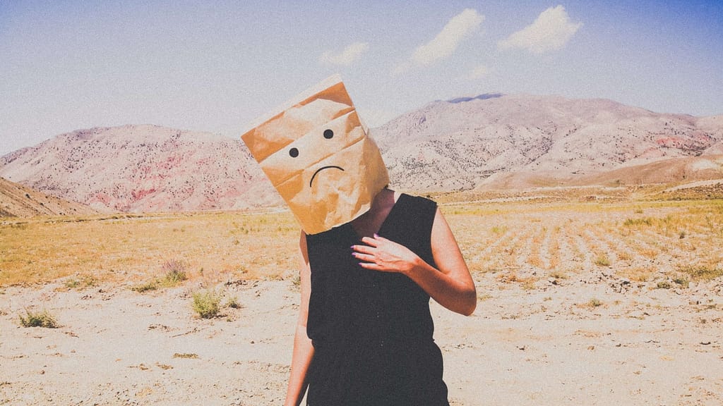 Image: A woman standing in a desert with a paper bag on her head, a frowning face drawn on the front. Symbolic of the emotions felt when we constantly say "I'll be happy when..."