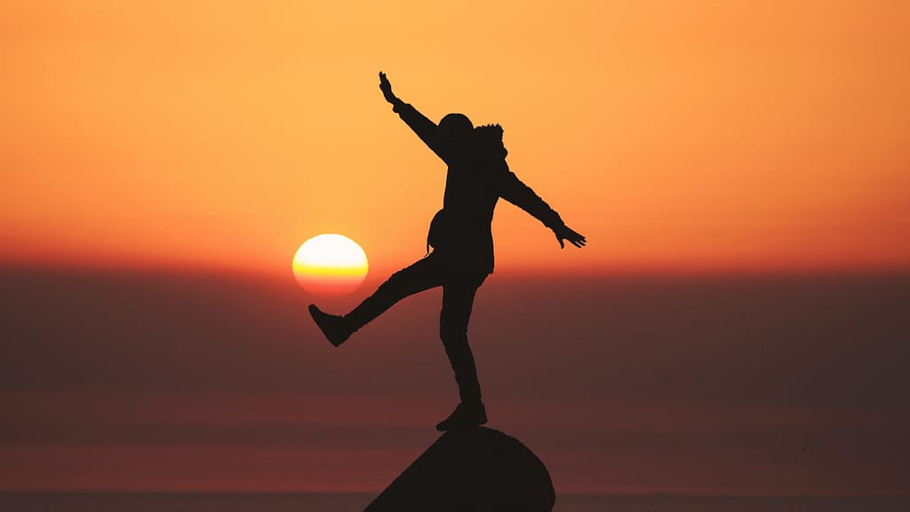 Image: An individual standing on a peak with one leg and both arms extended in front of an orange sunset, symbolizing that there is no such thing as peace of mind.