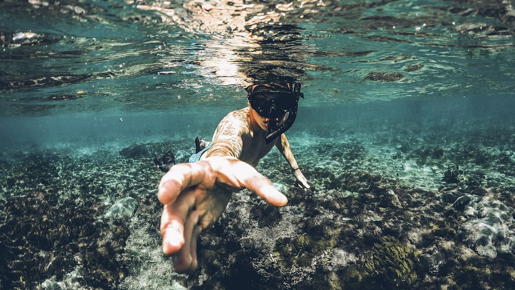Image: A man snorkeling above a coral reef in the ocean, reaching for the camera.