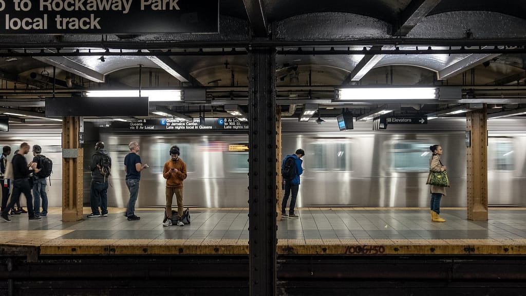 Image: People standing on a subway platform in NYC, a train rushing behind them. The place where Benny plays his trombone, after the accident where he lost all his memories.