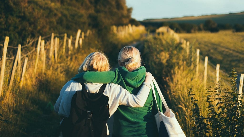 Image: Two women, arms slung around each other's shoulders, walking down a nature path. The two share their troubles with each other.
