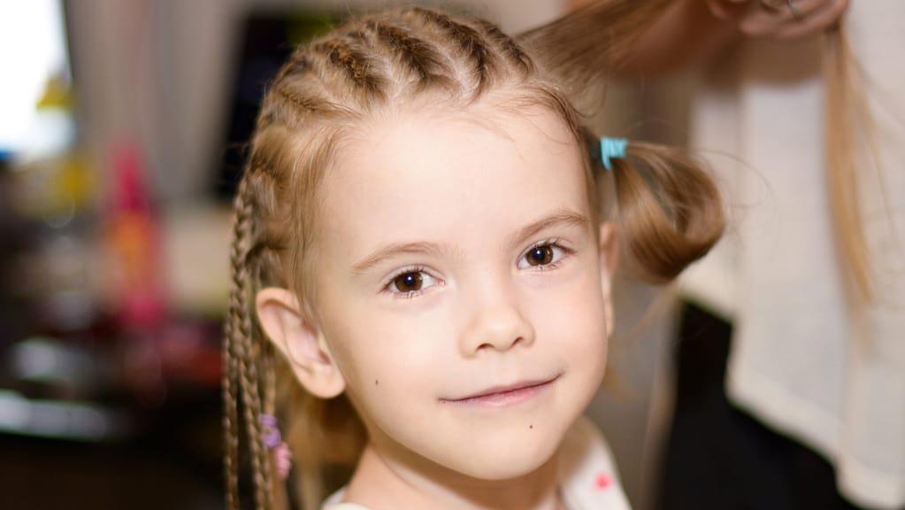 Image: a young girl looking at the camera, having her hair braided to improve self-esteem