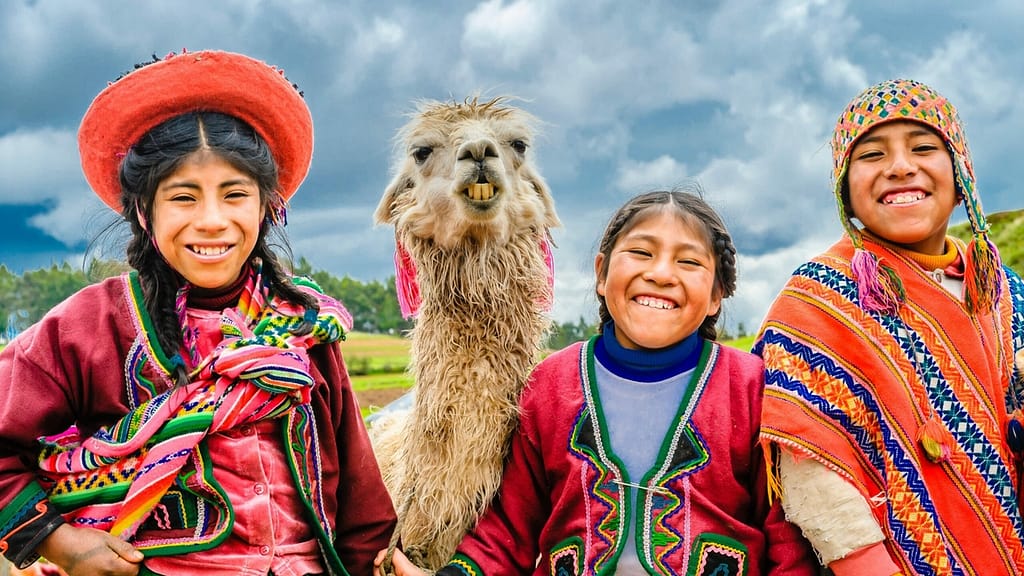 Image: Three Andean children standing next to an alpaca in Cusco, Peru, smiling.