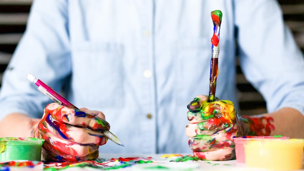 Image: A man sitting at a table painting, his hands absolutely covered in colored paint, representing the ability to let go of perfectionism. 