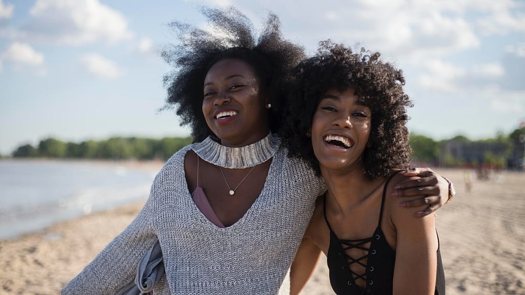 Image: Two black women on a beach, one with her arm slung around the other's shoulders, clearly demonstrating that this is one of their valued friendships.