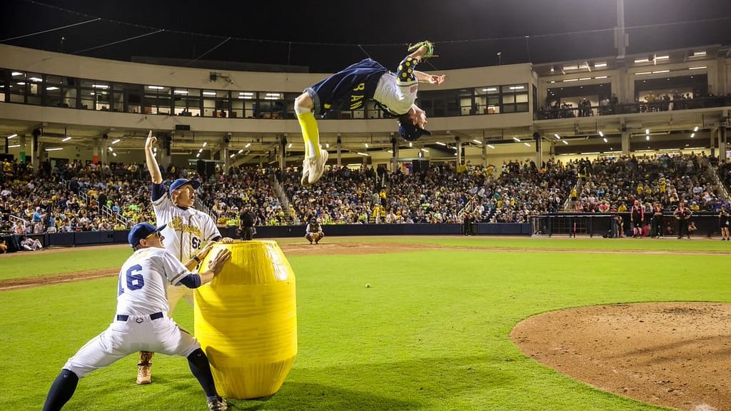 Image: A player for the Savannah Bananas doing a backflip off of a barrel in front of a cheering crowd.