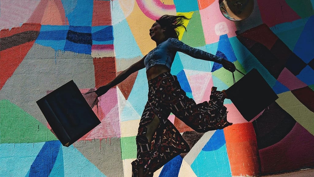 Image: A woman holding shopping bags full of things which she went to buy, dancing in front of a mural.