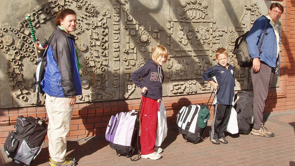 Image: kids standing in a line with their father wit their backpacks and suitcases behind them.