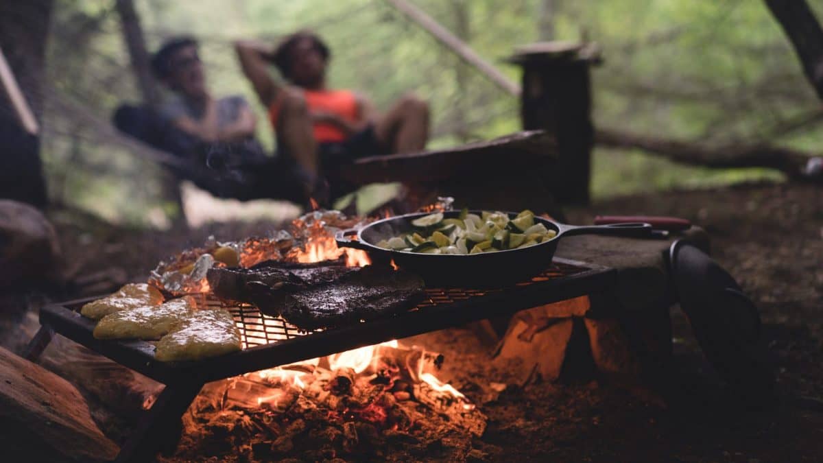 Image: Steak, potatoes, and vegetables cooking on a grate over a hot campfire.