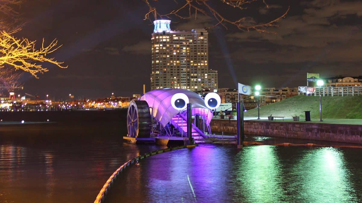 Image: Mr. Trash Wheel on the Baltimore River at night, lit up by purple lights with the Baltimore skyline in the background.