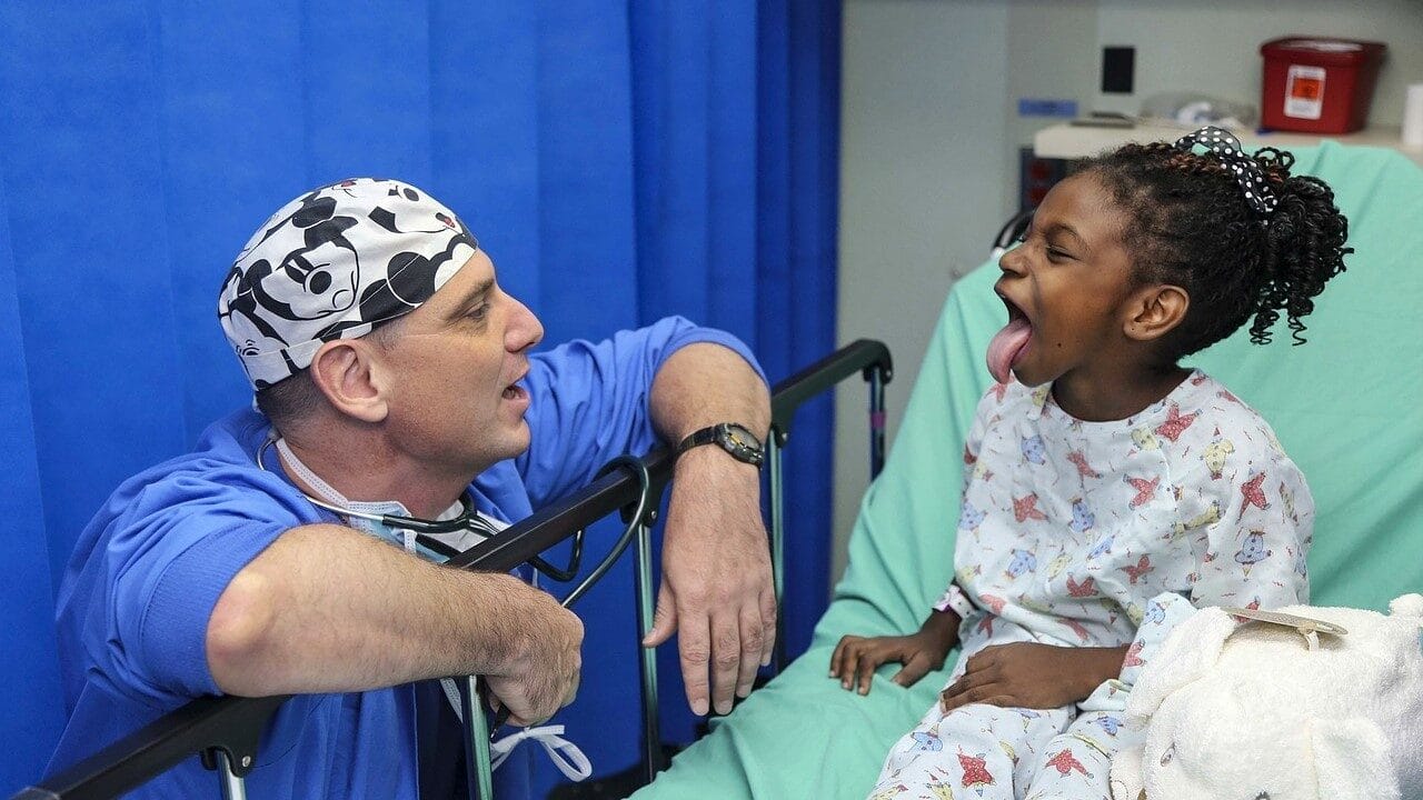 Image: Doctor sitting next to a hospital bed, wearing blue scrubs and a Mickey Mouse patterned head cap, interacting with a young girl who's sticking her tongue out. Feeling is joyful!