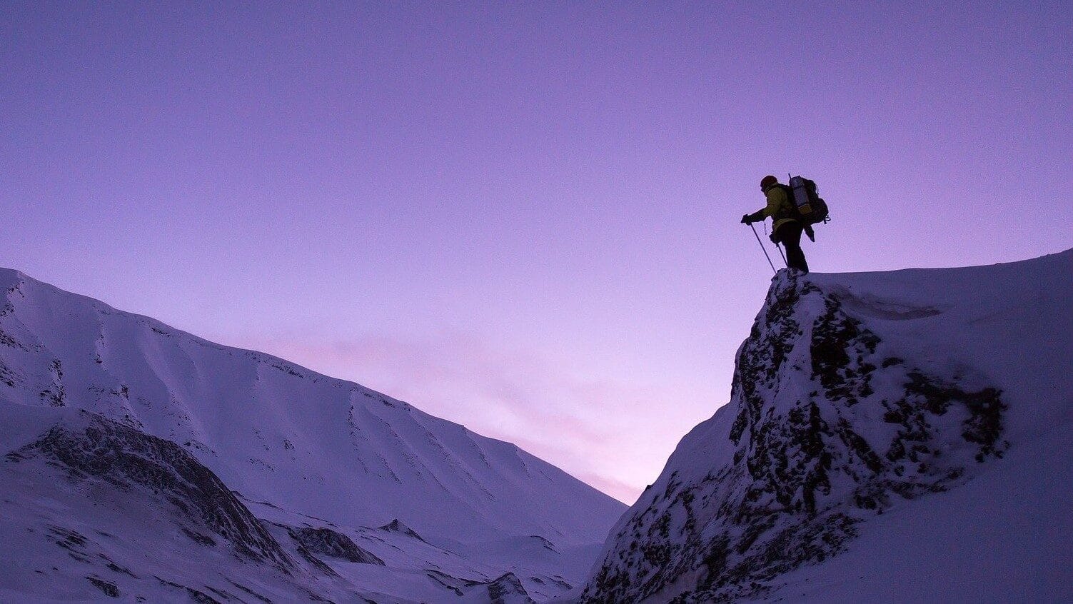 Image: Person with large backpack on standing atop of a snow covered cliff, staring off into the distance.
