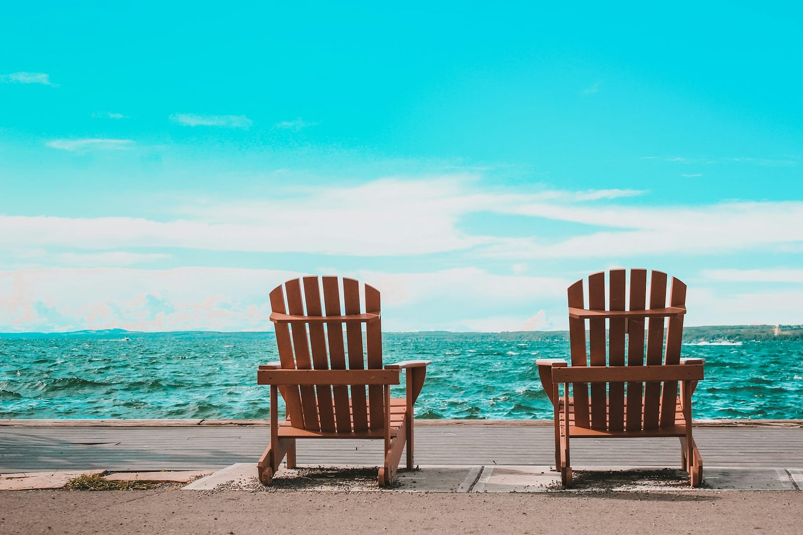 Image: Two empty wooden chairs overlooking a bright blue ocean and sky