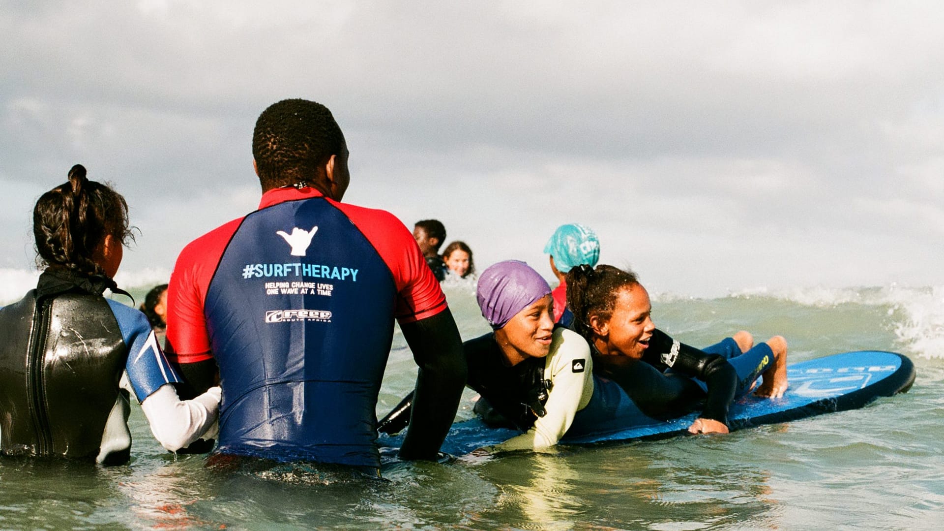 Image: two children in the ocean on a surfboard being held in place by a coach who sports a #SurfTherapy graphic on their shirt