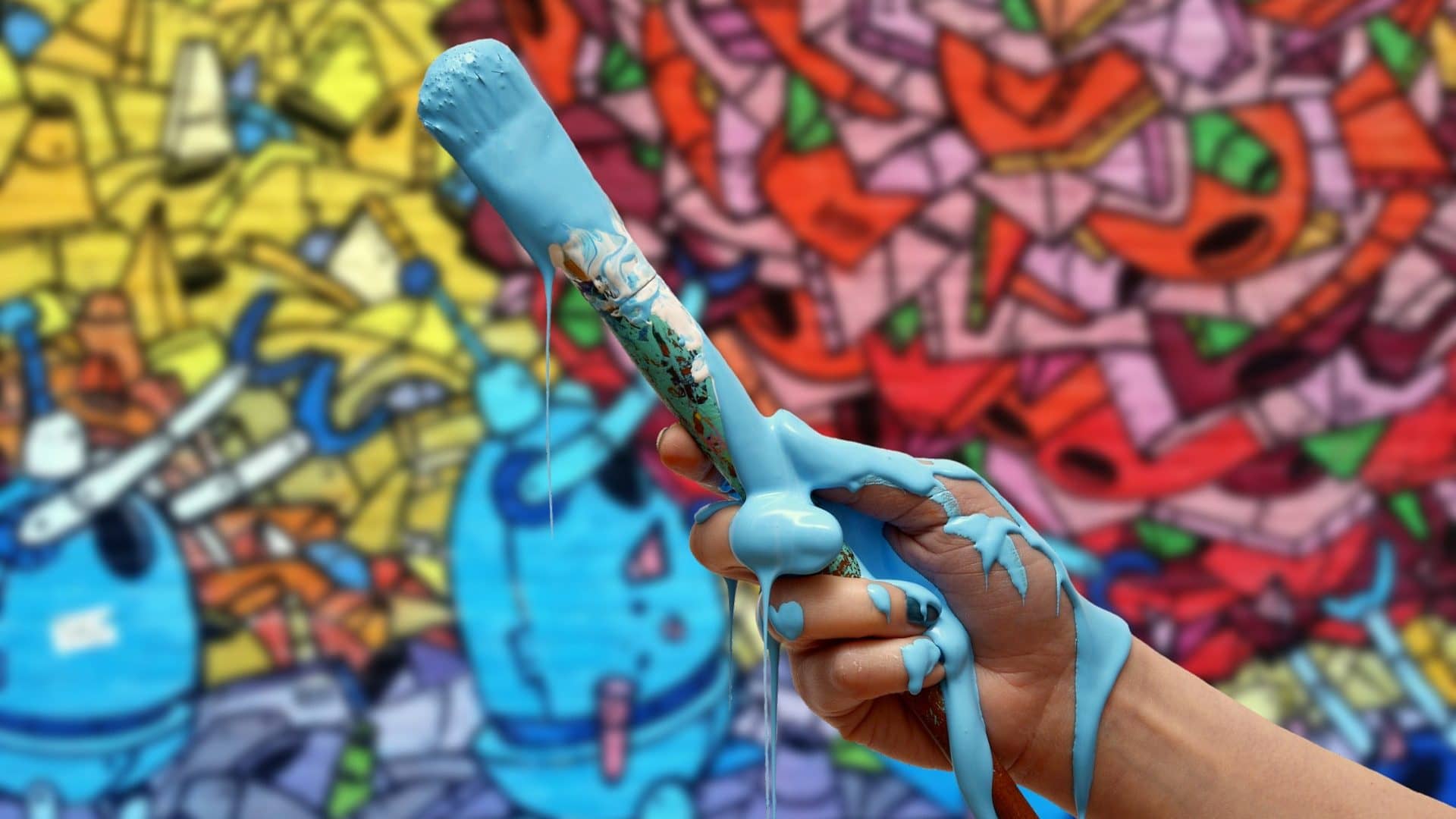 Image: Person holding a paintbrush covered in paint in front of a wall with graffiti on it