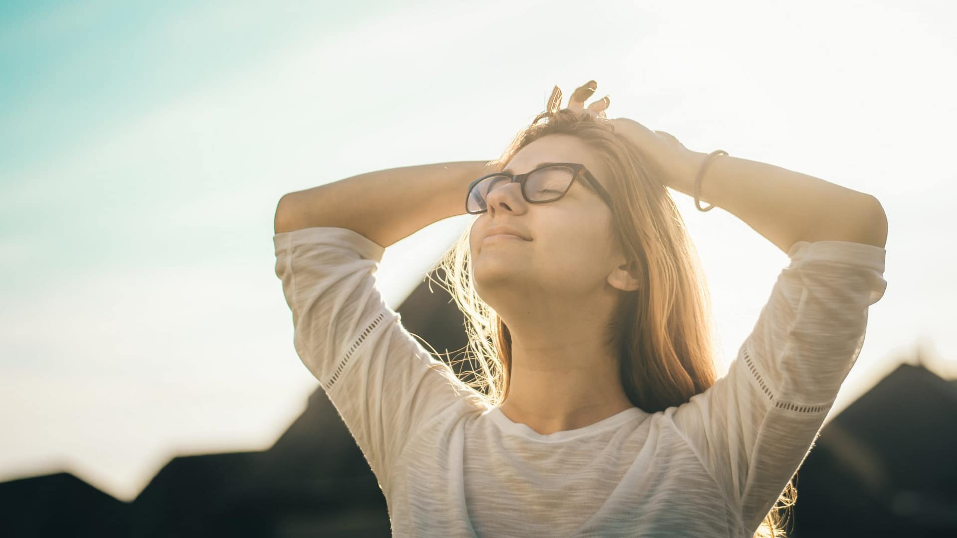 Image: A blonde woman wearing glasses, standing outside with her hands behind her head, with a look of ease and joy on her face