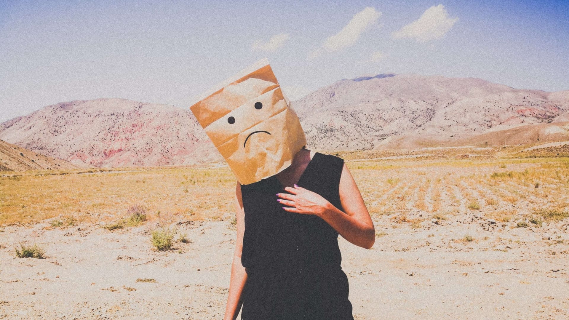 Image: A woman standing in a desert with a paper bag on her head, a frowning face drawn on the front. Symbolic of the emotions felt when we constantly say "I'll be happy when..."