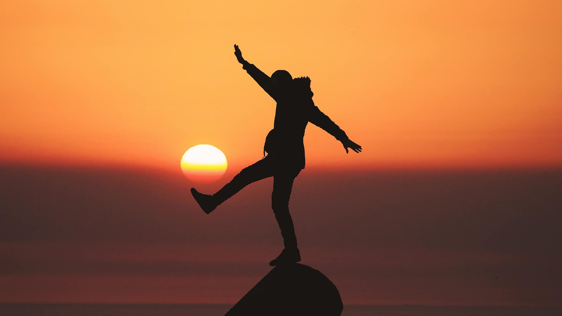Image: An individual standing on a peak with one leg and both arms extended in front of an orange sunset, symbolizing that there is no such thing as peace of mind.