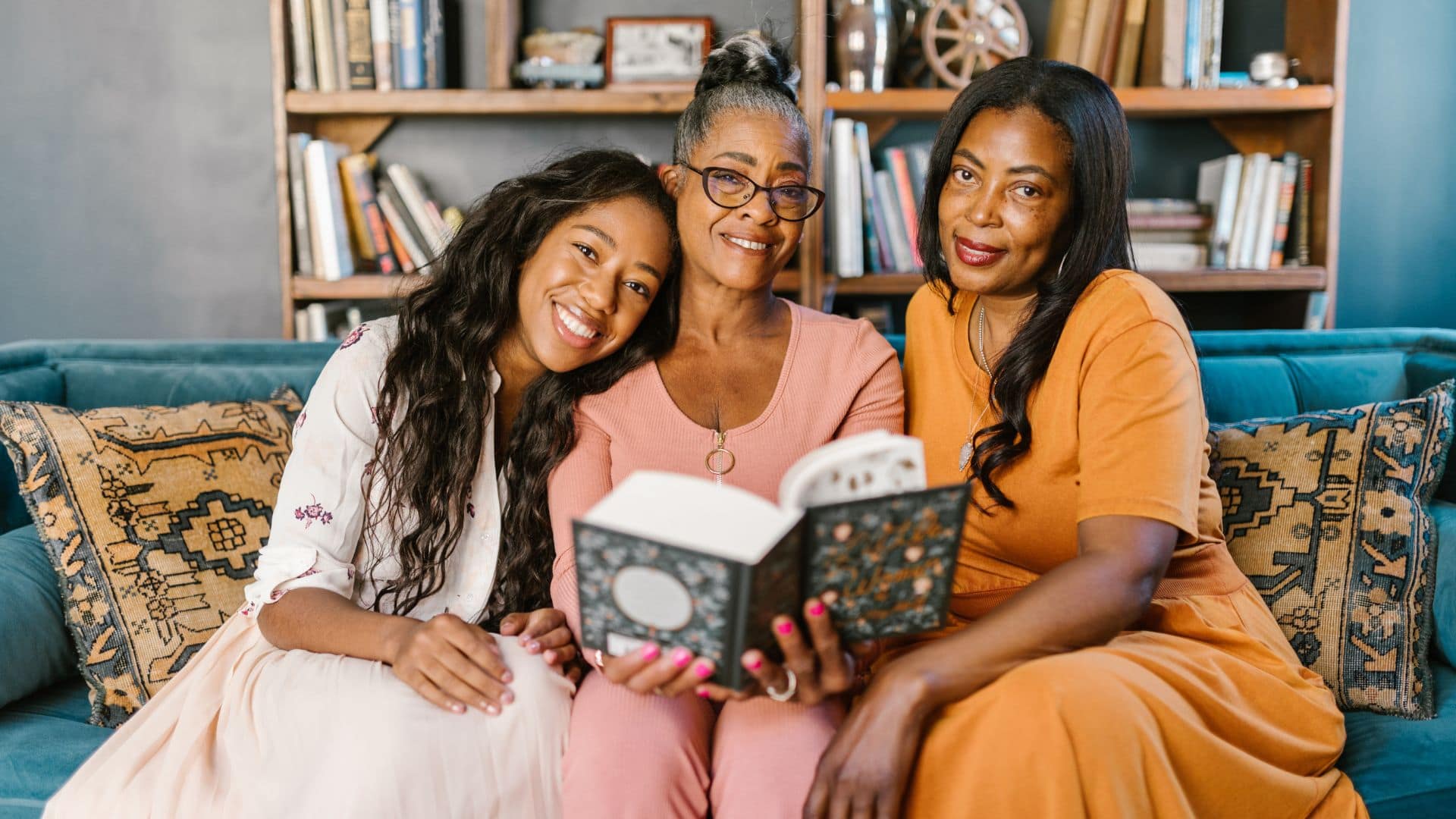 Image: a 3 generations of women sit together on a couch reading a book
