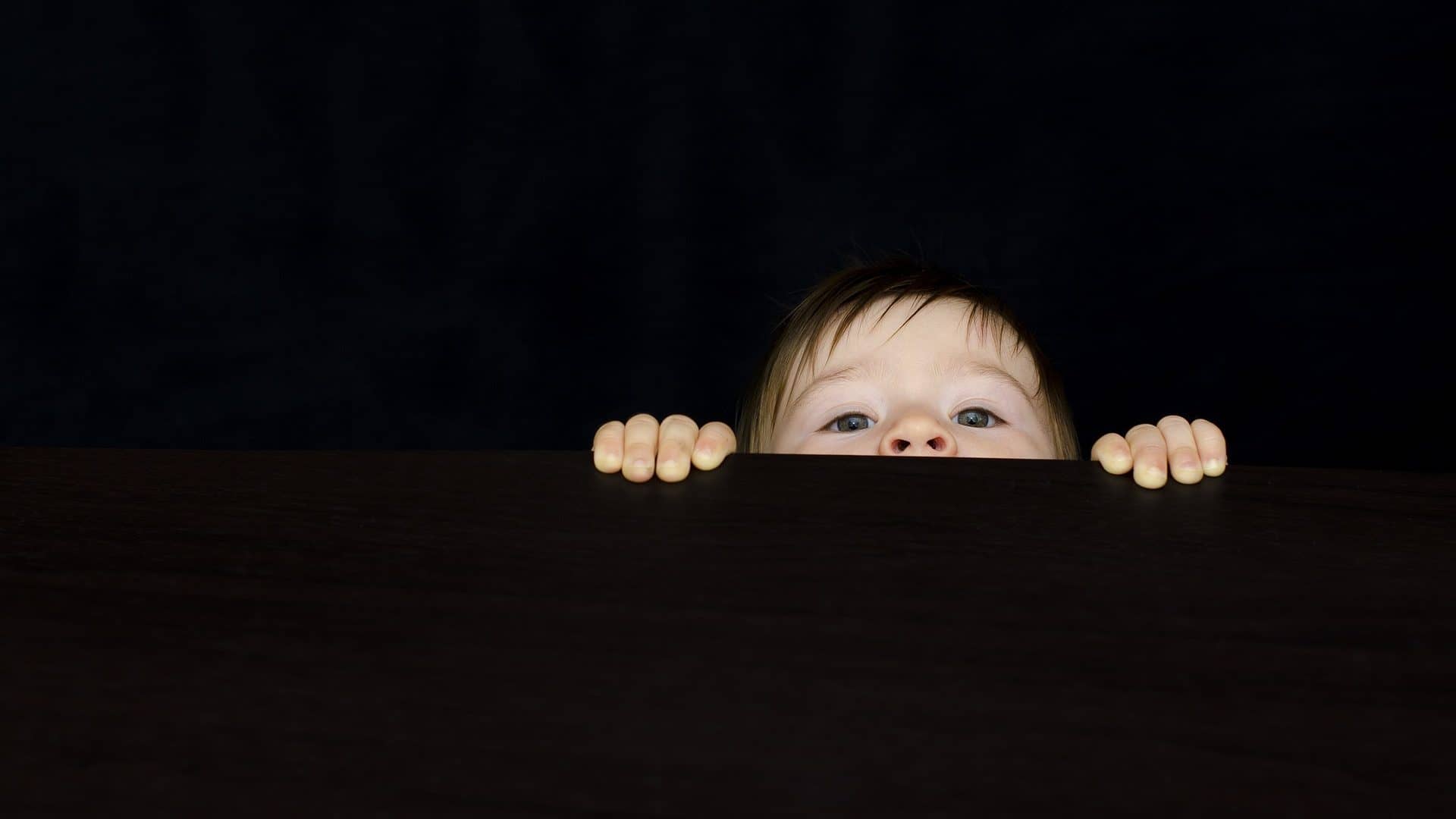 Image: A little boy peeking over a black wall, only eyes, nose and hands showing.