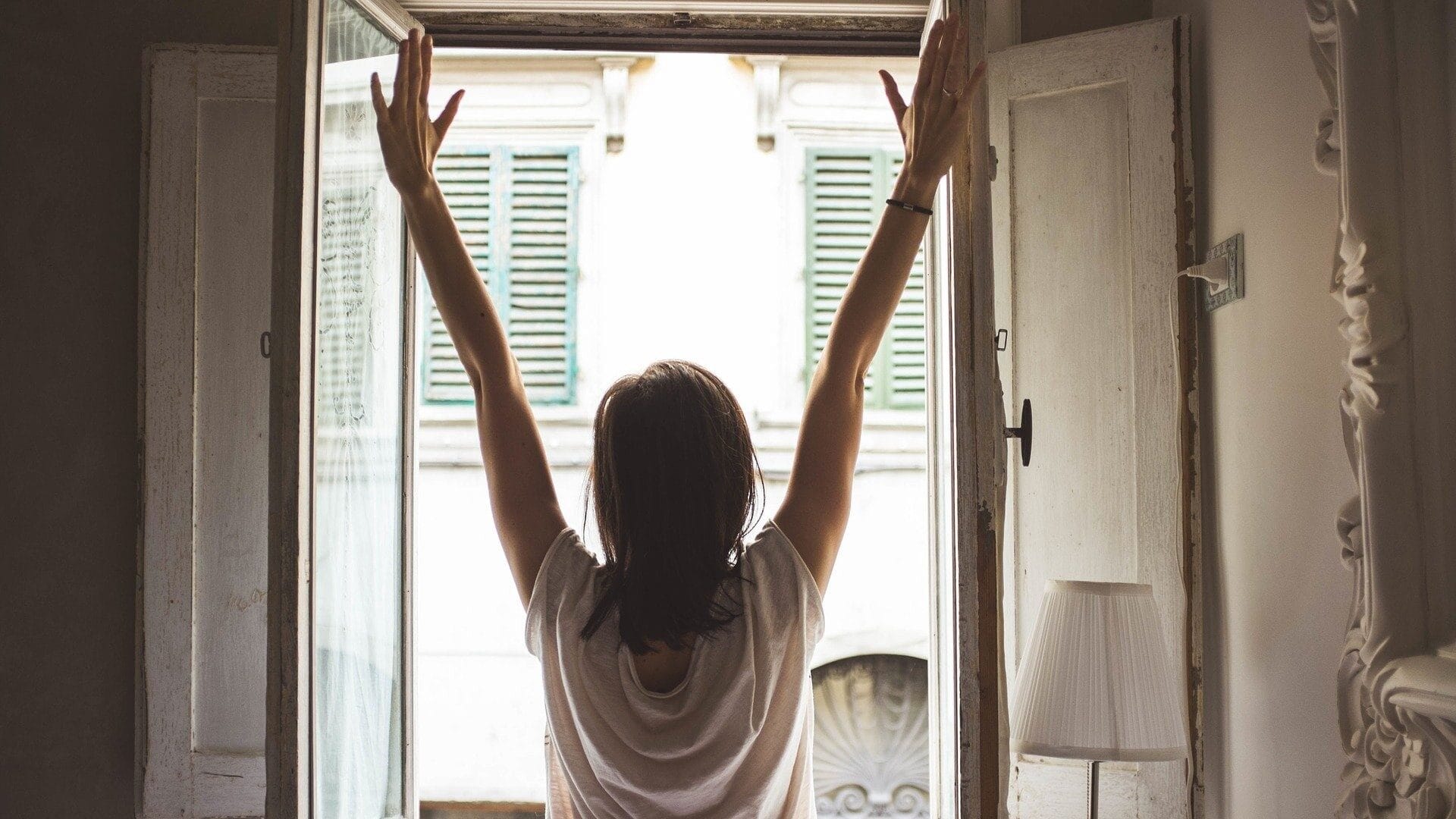 Image: Person raising their arms with joy to the morning sun at their window