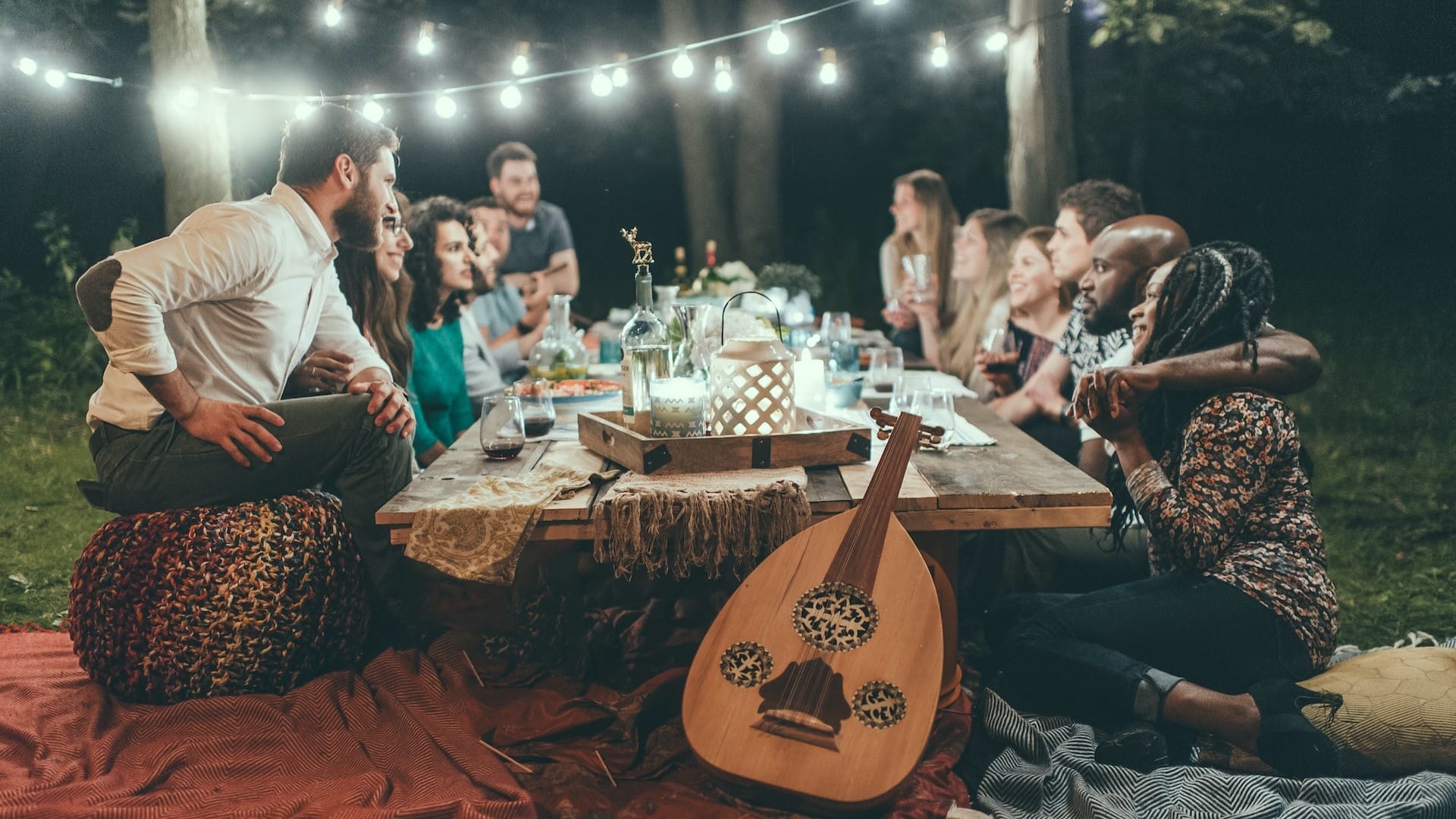 Image: A group of friends chatting over a table, with fairy lights, a guitar, and food. They are clearly reveling in the importance of human connection