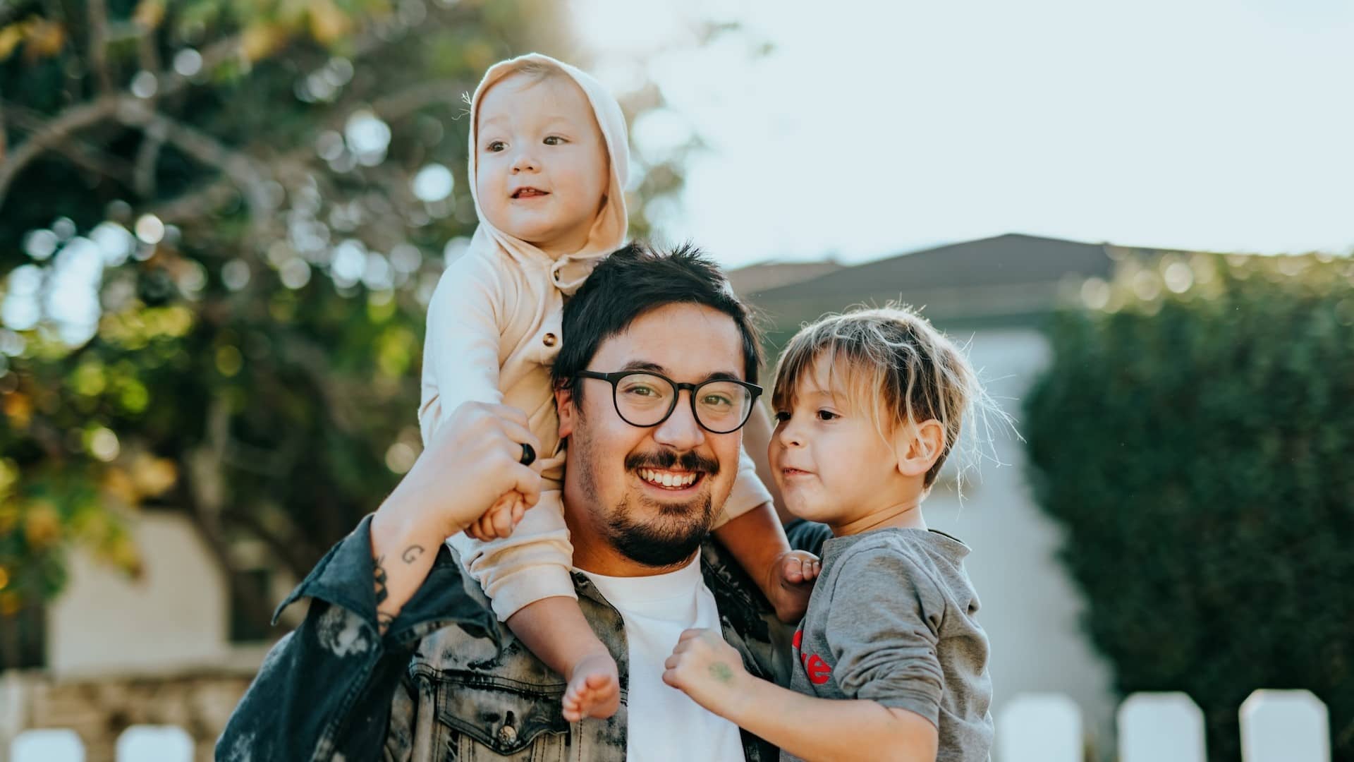 Image: A man with his two children, one child on his shoulders and the other in his arms, looking directly into the camera.