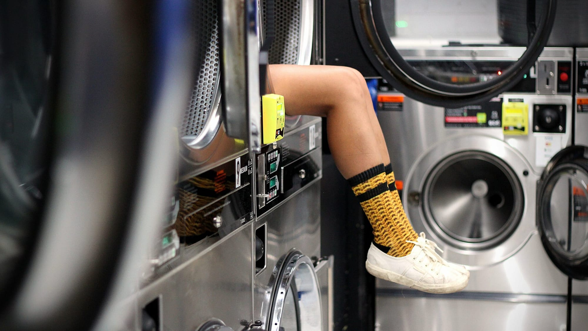 A pair of legs sticking out of a dryer, wearing long yellow socks and white shoes, at a laundromat.