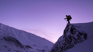 Image: Person with large backpack on standing atop of a snow covered cliff, staring off into the distance.
