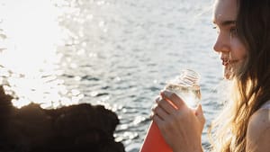 Image: A woman sitting by the sea, drinking water out of a water bottle.