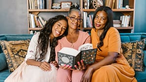 Image: a 3 generations of women sit together on a couch reading a book