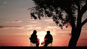 Image: two people sitting in chairs underneath a tree silhouetted against a sunset