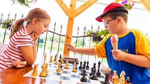 Image: two kids playing chess in a public park. On the left, a girl staring at the board, playing white. On the right, a boy holding an ice cream cone and making his move, playing black.
