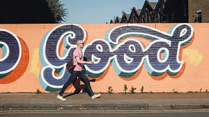 Image: A man walking down a side walk in front of an orange mural with blue writing that says "Good", using street wisdom practices.