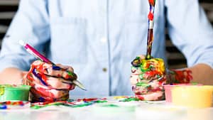 Image: a male artist sitting at a table, his hands covered in paint.