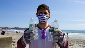 Image: A zero waste advocate taking park in a beach cleanup, holding two plastic water bottles that have been polluting the beach.