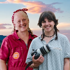 Beth White (left) and Zoë Brosky (right) of Habitat for Aviation's Women Build Planes stand against a sky with orange and purple clouds. White wears pink coveralls, and Brosky holds a professional camera.