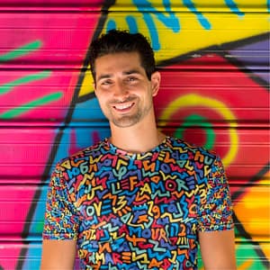 Artist Marco Santini stands smiling in front of one of his vibrantly colored 'One Love' murals, wearing a shirt with the same theming: the word love is spelled out in multiple colors against a black background