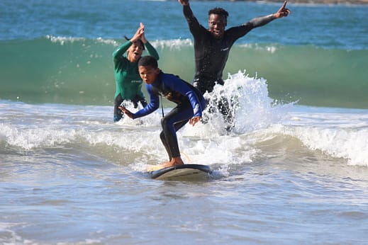 Image: A young person surfing on a small wave with two people cheering for them in the background