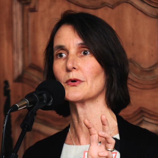 Liz Stookey Sunde, co founder of music to life, speaks into a large microphone. She is gesturing a grasp with one hand and holding a book in the other.