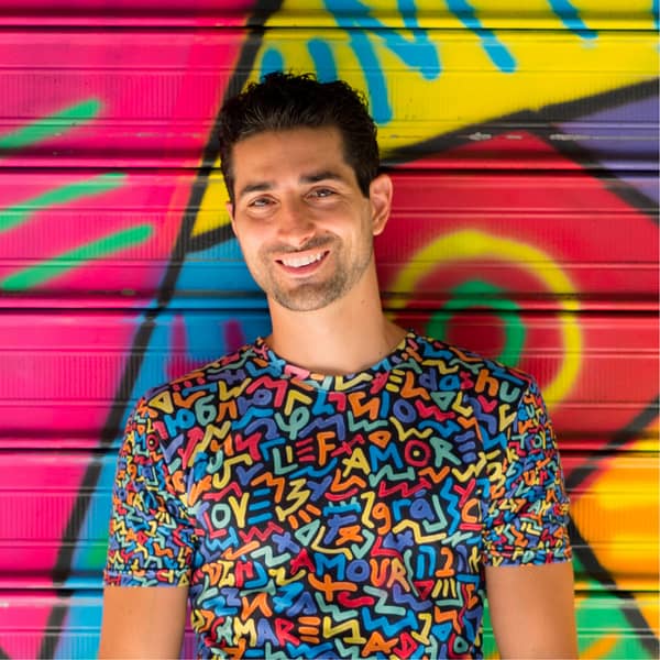 Artist Marco Santini stands smiling in front of one of his vibrantly colored 'One Love' murals, wearing a shirt with the same theming: the word love is spelled out in multiple colors against a black background