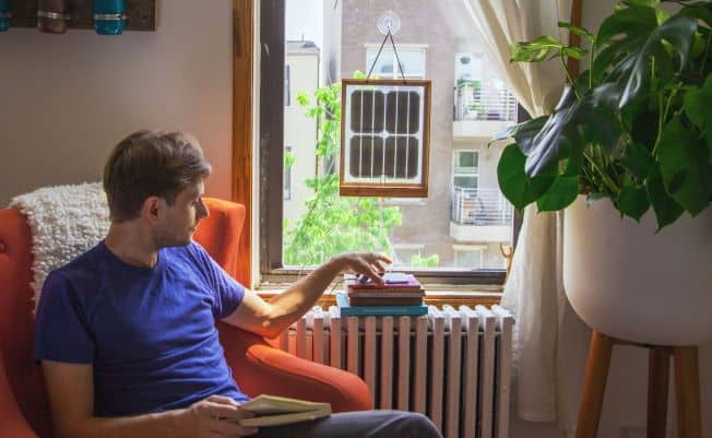 Image: Grouphug Solar Panel hanging in a window. Beside it, a man sits, looking out the window.