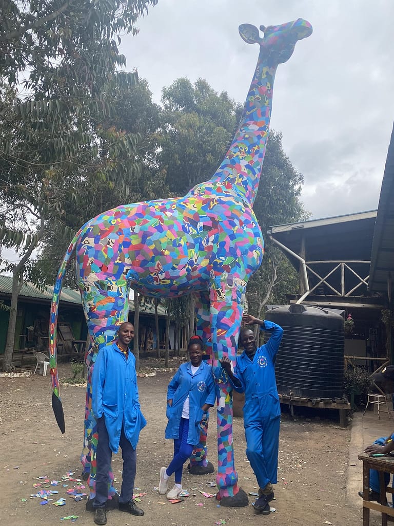 Image: Three Ocean Sole artisans standing beneath their life-size giraffe sculpture.