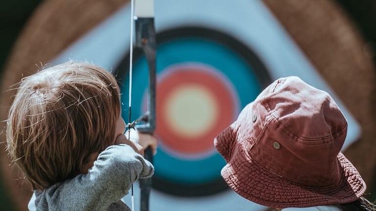 Image: Two children looking towards an archery target, the one on the left holding a bow and aiming at it.