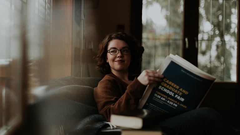 Image: A young woman with brown, curly hair sitting on a couch with a book in hand, looking out the window and contemplating her daily routine