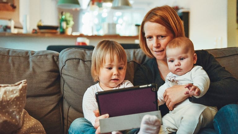 Image: A mom sitting on the couch with her two young kids, looking at a computer to volunteer from home.