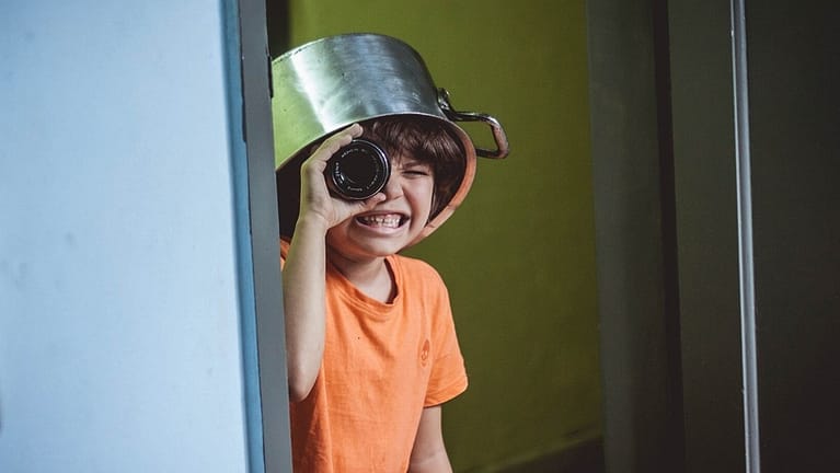 Image: child wearing a kitchen pot on their head, peeking around the corner with a camera lens as a telescope