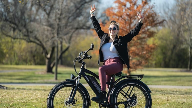 Image: A woman sat astride one of her local library's ebikes, holding her hands up in the air happily.